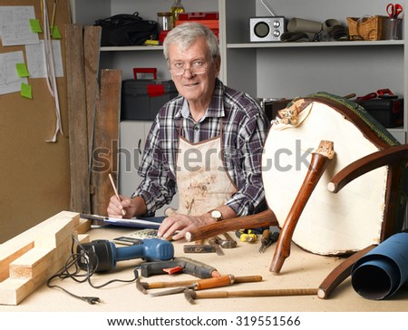 Portrait of retired manual worker working at his workshop. Senior carpenter writing on clipboard while sitting at desk and looking at camera.
