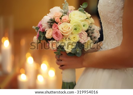 Closeup of young bride in white dress holding one beautiful fresh wedding bouquet of rose flowers pastel colors with blazing candles on blur warm light background, horizontal picture 
