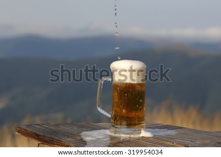 One glass mug pouring with lager or porter delicious frothy beer on wooden table top sunny day outdoor on natural with mountain hills and yellow dry grass background, horizontal picture