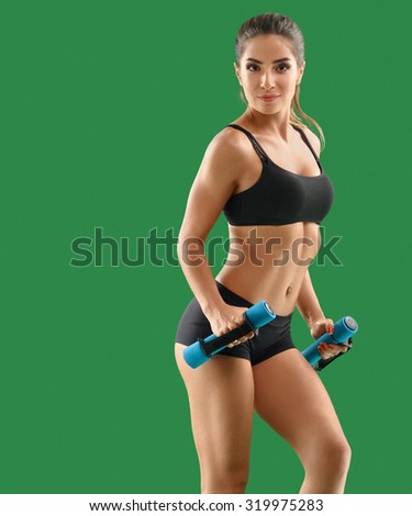 Slim, pretty young woman, sportswoman with beautiful athletic body doing exercises with dumbbells, looking in camera, in black sports wear on a green background. Hands and legs are bent; side view.