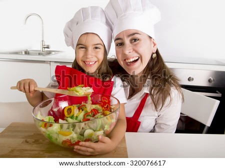 happy young mother and little daughter at home kitchen having fun preparing salad bowl wearing apron and cook hat playing together in healthy vegetables nutrition education concept