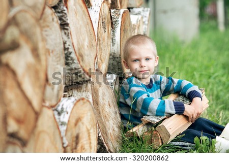 a small village boy blond 5 years sitting on the grass near a woodpile in rubber boots and a sweater with stripes from birch wood impaled in his hands and smiling