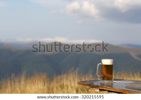 One glass mug with lager or porter tasty frothy beer on wooden table top sunny day outdoor on natural with mountain hills and yellow dry grass and cloudy sky background copyspace, horizontal picture