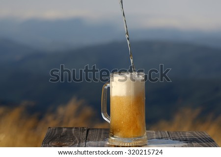 One glass mug pouring with lager or porter delicious frothy beer on wooden table top sunny day outdoor on natural with mountain hills and yellow dry grass background, horizontal picture
