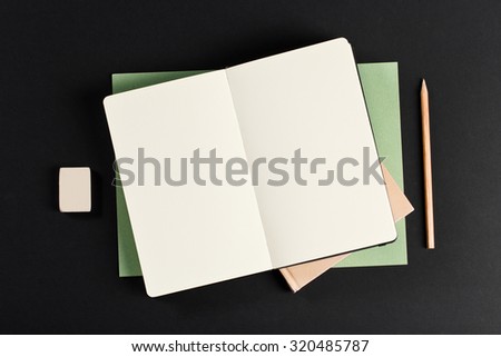Open blank notepad with empty white pages laying above book with pencil and eraser on black background. ideal for text