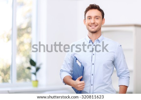 Ready to sale. Pleasant cheerful handsome realtor holding folder and expressing positivity while standing near window