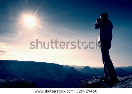 Professional photographer takes photos with big camera on peak of rock. Dreamy misty landscape, hot Sun above
