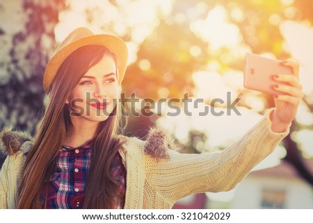 Beautiful teenage girl in beige hat and sweater taking a selfie on smartphone outdoors in park on sunny autumn day. Cute young woman photographing herself. Retouched, horizontal, vibrant colors.