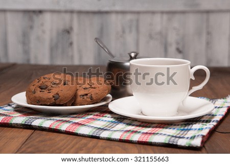 Picture of stilllife with a cup, sugar bowl and saucer with cookies represented on table-top cloth on wooden table.