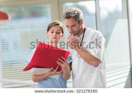 A nice grey hair doctor with beard and a nurse are examining a patient file He is wearing his white coat, his stethoscope around his neck. They are standing in front of hospital room with glass walls