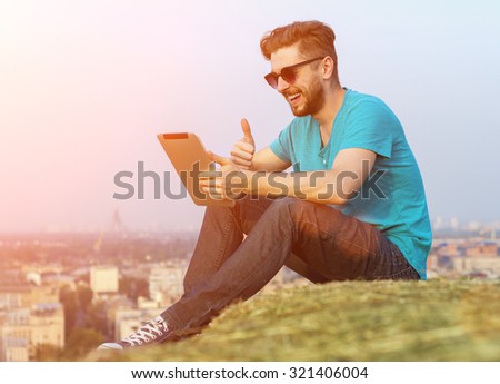 Man Sitting On A Mountain On The Background Of The City Is Holding A Computer Tablet And Laughs