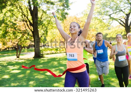 fitness, sport, victory, success and healthy lifestyle concept - happy woman winning race and coming first to finish red ribbon over group of sportsmen running marathon with badge numbers outdoors Royalty-Free Stock Photo #321495263