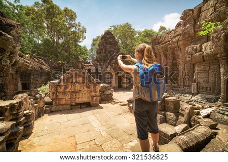 Young female tourist with blue backpack ands smartphone taking picture of the unique bas-relief among mysterious ruins of the ancient Preah Khan temple in Angkor. Siem Reap, Cambodia.