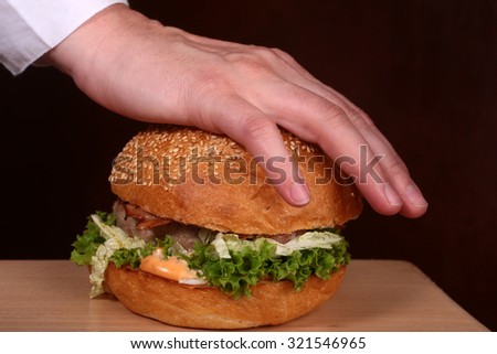 Big tasty appetizing fresh burger of green lettuce red tomato cheese and bacon slice meat cutlet and white bread bun with sesame seeds pressed by human hand on wooden table top, horizontal picture