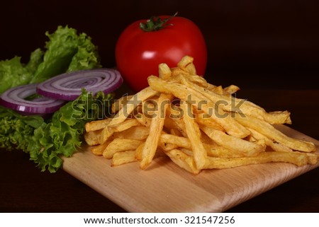 Closeup of fried fresh tasty potato chips sticks golden color lying in heap near red tomato violet onion and green lettuce on wooden background, horizontal picture