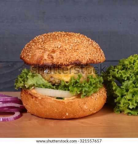 One big tasty appetizing fresh burger of green lettuce red tomato cheese bacon slice meat cutlet violet onion and white bread bun with sesame seeds on wooden table closeup, square picture