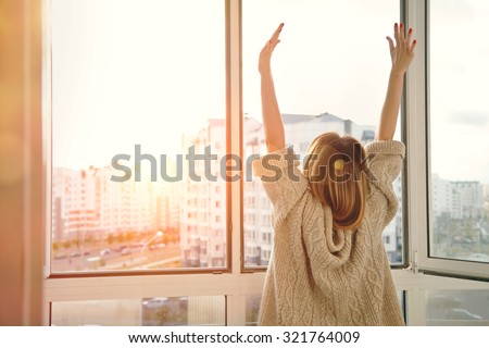 Woman near window raising hands facing the sunrise at morning Royalty-Free Stock Photo #321764009
