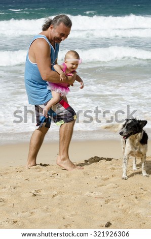 European family. Father dad man with long hair style and daughter little happy cute pretty baby infant girl in pink dress walking on the beach near sea and looking the dog on the background of waves