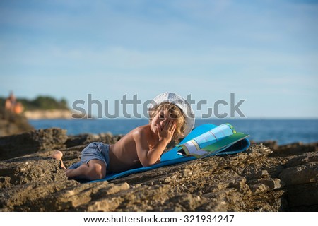 Cute little boy with a cap lying on his stomach looking at his picture book on the beach. Some negative space around.
