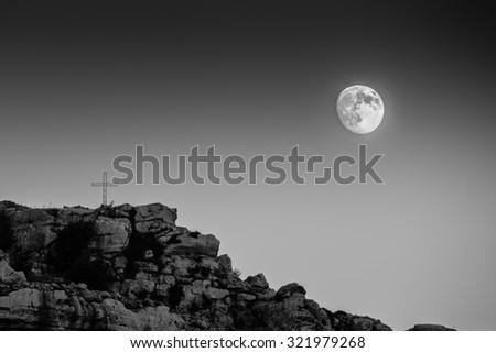 Black and white picture of a nearly full moon rising behind a cross on top of one of the surrounding hills of the town of Jezzine, Lebanon.