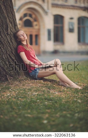 Young pretty student woman is sitting near the tree with a book in her hands and dreaming with joyfull and relaxed expression in the university campus. Love to study concept. 