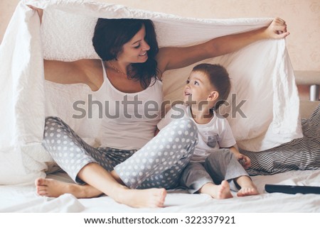 Young mother with her 2 years old little son dressed in pajamas are relaxing and playing in the bed at the weekend together, lazy morning, warm and cozy scene. Pastel colors, selective focus. Royalty-Free Stock Photo #322337921