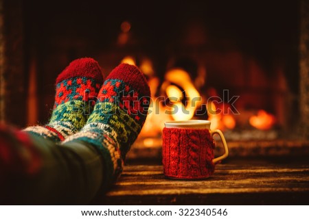 Feet in woollen socks by the Christmas fireplace. Woman relaxes by warm fire with a cup of hot drink and warming up her feet in woollen socks. Close up on feet. Winter and Christmas holidays concept. Royalty-Free Stock Photo #322340546