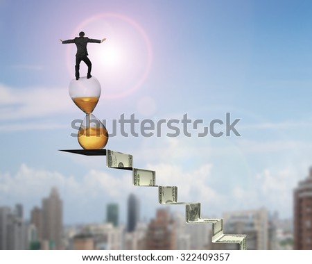 Businessman balancing hourglass on top of money stairs, with sun sky cityscape background.
