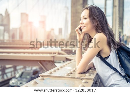 Young pretty woman talking at cellular phone on Brooklyn Bridge - Mixed race woman having a conversation at smartphone, city at sunset in the background Royalty-Free Stock Photo #322858019