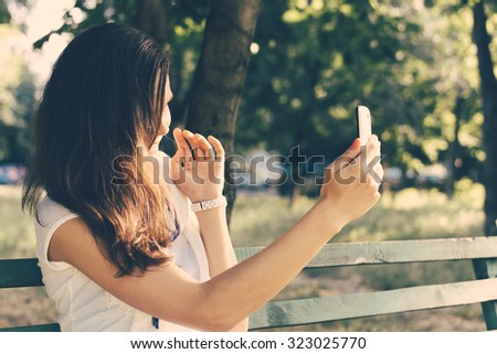 Young woman sitting on a park bench and photographed on a mobile phone. Leisure at outdoors on a sunny day in the summer. Retro colors.
