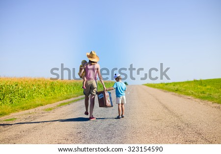 Picture of young woman carrying baby and suitcase walking away on road with little boy. Family travelling by foot on sunny summer countryside background.