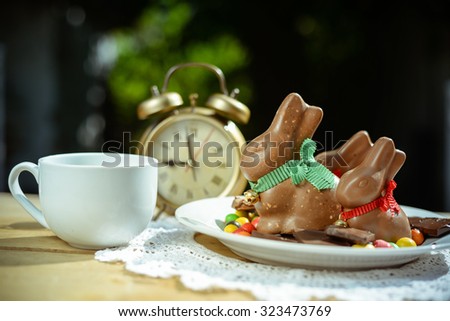 Picture of three big chocolate bunnies on white plate. Colorful candies and cup beside retro alarm clock on blurred indoor background.