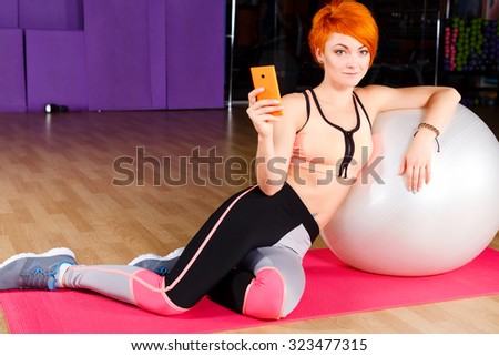 Young pretty woman, with short haircut, wearing in top and leggings, sitting on pink yoga mat with fitball and smart phone, in the gym, full body