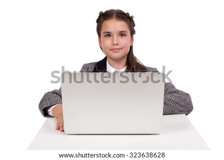 Beautiful school girl in a business suit sitting at a table in front of a laptop and a smile, looking into the camera. Photo from the depth of field