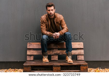Confident and handsome. Handsome young man sitting on the wooden pallet and looking at camera with grey wall in the background and orange fallen leaves on the floor  Royalty-Free Stock Photo #323733749