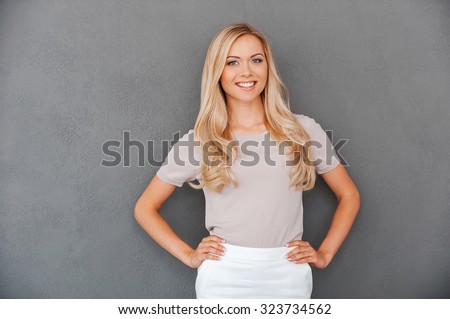Confident in her abilities. Smiling young blond hair woman holding hands on hips and looking at camera while standing against grey background