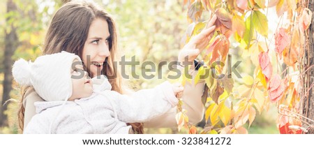 Pretty mother with her baby outdoor at autumn park, wearing a coat and touching the leaves and trees. At the cold time you need dressing your baby warm - hat, jacket and pants