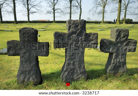 Three stone crosses, made of rough-hewn granite, marking the graves of unknown soldiers in a German military cemetery. A single poppy at the base. Two figures can be seen in the background.