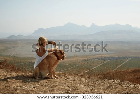 Young woman with dog on a sunny day sitting in high mountains and take a picture on smartphone.
