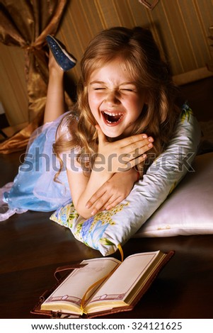 Little smiling girl in a blue dress lies on the floor in room, beside her is a book
