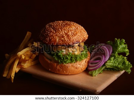 One big tasty appetizing fresh burger of green lettuce cheese bacon slice meat cutlet and white bread bun with sesame seeds and potato chips on wooden table and potato chips, horizontal picture