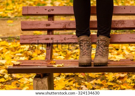 Girl in boots standing on a red bench with yellow birch leaves in the background. Autumn picture in a park. Cute fall image.