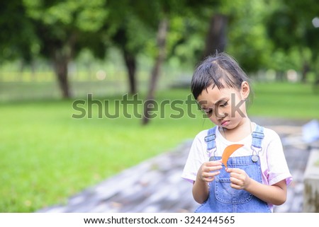 Little Asian girl on thinking pose; Kid look at yellow leaf and think; Blur green park background