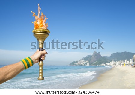 Hand of an athlete wearing Brazil colors sweatband holding sport torch on Ipanema Beach with Two Brothers Mountain on the skyline of Rio de Janeiro, Brazil