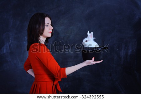 Teacher of of Magic. Young beautiful girl in a red dress holds a black illusionist hat with a white rabbit inside it with a blackboard background. Alice in Wonderland