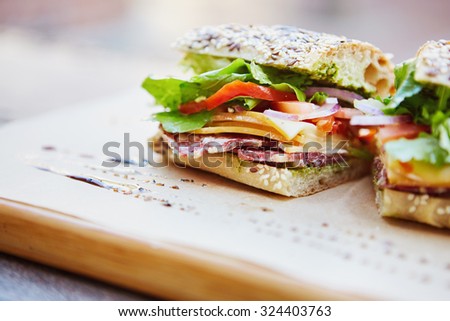 Healthy sandwich made of a fresh seeded roll, cut in half to display tasty ingredients of salami, tomato, lettuce and chess, presented on a wooden board Royalty-Free Stock Photo #324403763
