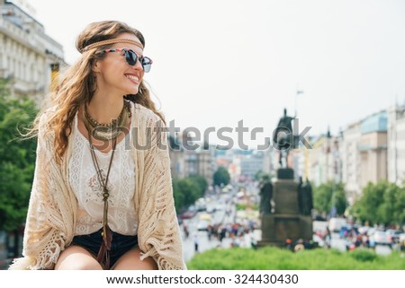 Smiling trendy hippie brunette woman tourist relaxing on stone parapet on Wenceslas Square in Prague. In the background Saint Wenceslas statue in Prague. Tourism travel concept.