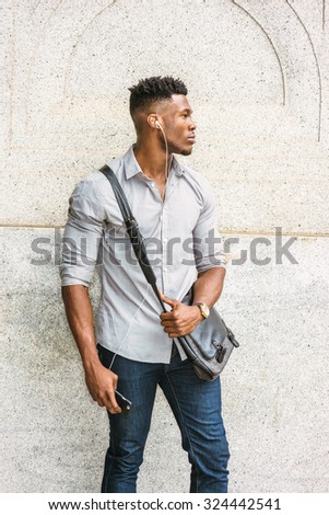 Man on the way going to work. Wearing gray shirt, jeans, wristwatch, carrying shoulder leather bag, an African American guy standing by wall on street, listening music with earphone and cell phone.
