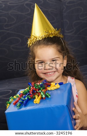 Hispanic girl wearing party hat holding birthday present smiling and  looking at viewer.