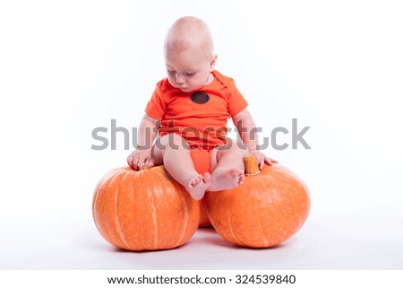 Beautiful baby in orange t-shirt on a white background sitting on a pumpkin and looking at the camera smiling, picture with depth of field